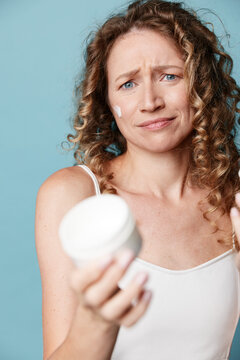 Middle aged woman with natural skin holds a cream jar and looks puzzled at the camera. Studio shot on a blue background. Concept for skincare, beauty routine, or product testing.