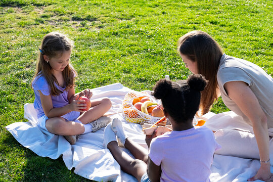 Family members sitting on a blanket on green grass, sharing healthy snacks during a park picnic