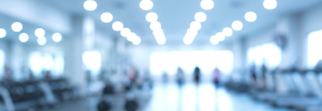A blurred image of a modern gym interior filled with exercise equipment and bright ambient lighting. The spacious area suggests an inviting workout environment.