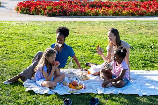 Group of diverse women and children sitting on a blanket, eating snacks outdoors