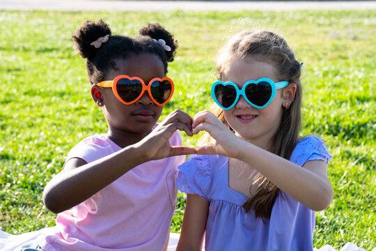 Two young girls wearing heart sunglasses creating a heart shape with their hands outdoors
