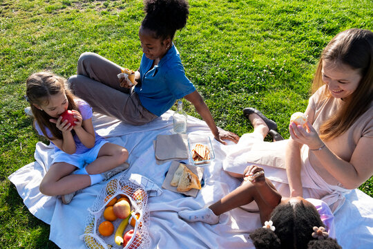 Four friends and family members relaxing on a blanket, eating snacks and fruit in a park
