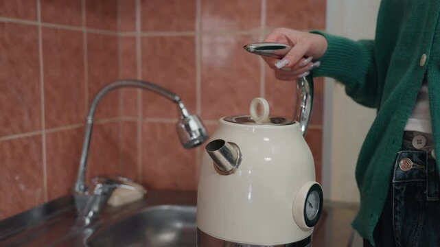 Young woman filling kettle in new apartment kitchen, green sweater hands under tap, vintage white kettle on counter, tiled backsplash and stainless sink visible, gentle morning light, subtle steam