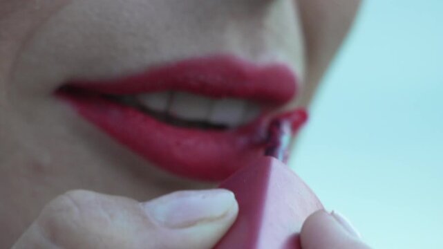 Vibrant red lipstick application on a woman, applying cosmetic color with a precision brush indoors. Extreme macro close-up shot.