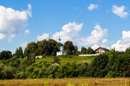 Picturesque white orthodox church with a green dome nestled among lush trees on a scenic grassy hill near residential houses under a bright blue summer sky with fluffy clouds. Kaluga oblast, Russia