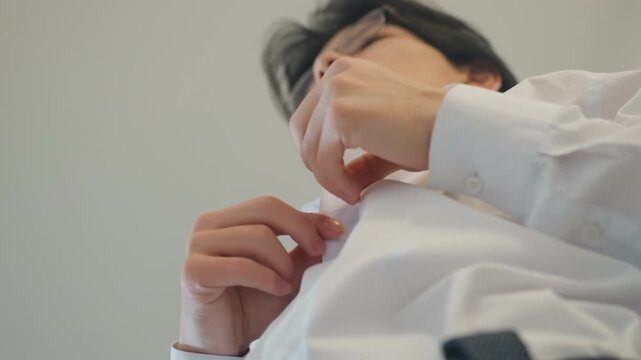 Young man buttoning white shirt in lowangle closeup, steady hands adjusting cuff and collar, soft morning light, minimalist wall background, getting ready as junior executive before work, precise