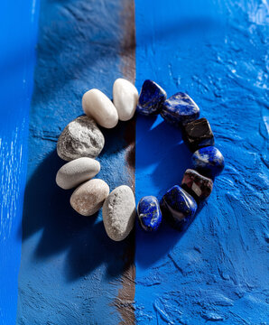 Circle of smooth stones and polished blue gems on blue textured background. Contrast between natural river rocks and precious minerals. Concept of balance, harmony and contrast.