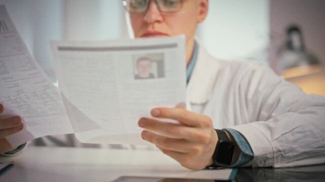 Doctor reviews a patient's documents, scans the medical card and checks the insurance policy while seated in his white office at the clinic, showing careful record-keeping in a professional workplace