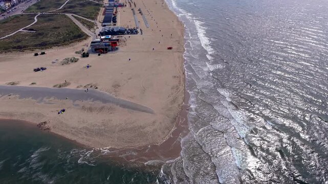 Aerial view video clip of Katwijk aan Zee sandy beach, Oude Rijn river mouth outlet, beachfront pavilions, glinting North Sea waves, and coastal dunes in South Holland, Netherlands