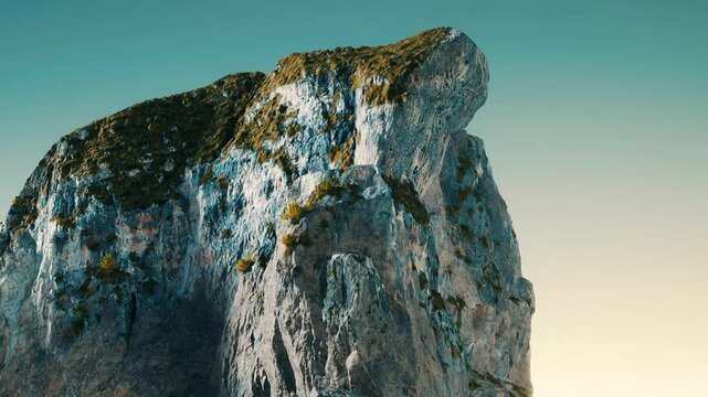 A large rock formation stands tall with green vegetation on its surface. The sky is clear during the time of sunset. The colors of the rock and sky merge peacefully.