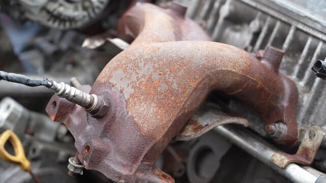 Rusty car exhaust manifold with oxygen sensor on a salvaged vehicle engine block. Close-up of a corroded automotive exhaust manifold with an attached oxygen sensor in a scrap yard.
