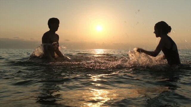 Happy teenage siblings splash water at each other in the sea at sunset in slow motion. Carefree youth, water play, summer vacation and unforgettable moments together