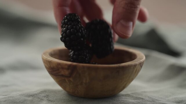 Slow motion man hand put fresh ripe blackberry in a small wooden bowl