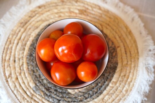 Top view of a bowl filled with ripe, red tomatoes placed on a decorative woven placemat. The vibrant tomatoes contrast with the textured background, highlighting natural produce