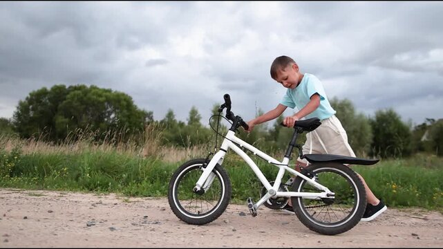 Boy walks beside white bicycle along rural gravel road. Side view shows cloudy fields trees and child learning independence during outdoor family time.