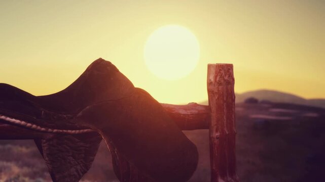 A saddle rests on a wooden fence in an open field. The sun sets in the background, casting an orange glow. Distant mountains provide a backdrop to the scene.