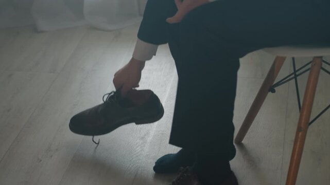 Man sitting in socks removing shoes, hands untying laces and easing foot out of brown leather, wooden chair and laminate floor, soft window light, slow intimate mood of transition after commute,