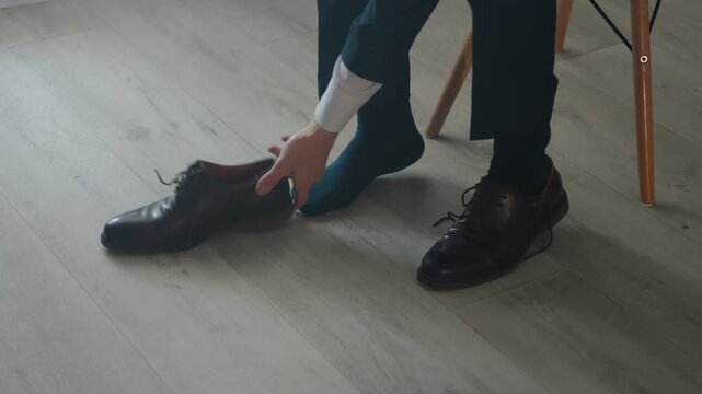 White businessman removing dress shoes on hardwood floor, seated on chair, untying leather brogues and placing shoes aside frames show executive untying after commute, groom preparing footwear,