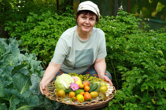  Smiling woman holding a basket filled with fresh, organic vegetables including tomatoes, cabbage, and cucumbers. Surrounded by lush greenery, showcasing homegrown produce joyfully.