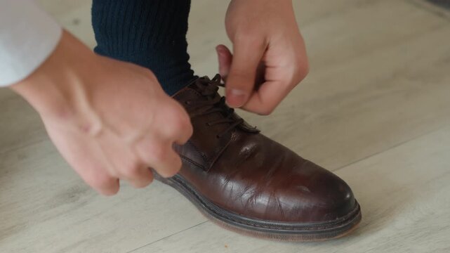 Close up brown leather shoe being laced man prepares for calm morning walk on wooden floor, navy socks, steady hands, vintage brogue details, warm natural light, intimate domestic atmosphere