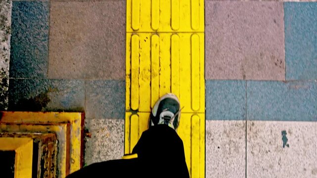 Point of view shot of a person walking forward along a bright yellow tactile paving path on a textured tile sidewalk during daytime.