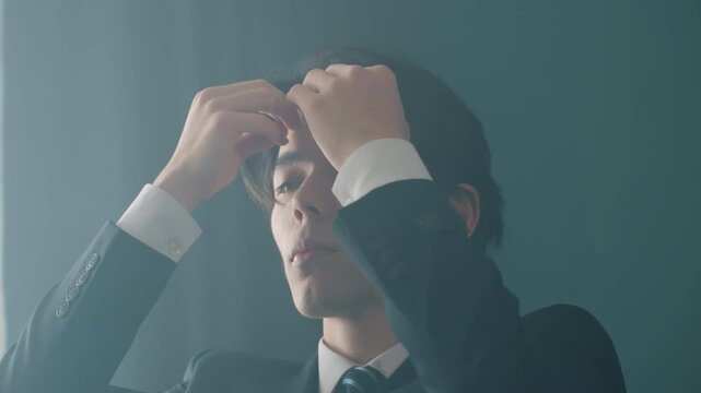 Asian businessman adjusting hair before meeting in moody office light wearing dark suit and tie, executive grooming ritual preparing for interview or pitch, confident composed expression, hands