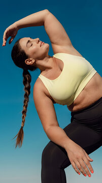Low angle view of peaceful plus size woman in light yellow sports bra and black leggings stretching side body with eyes closed against clear blue sky background, body positivity and outdoor yoga conce