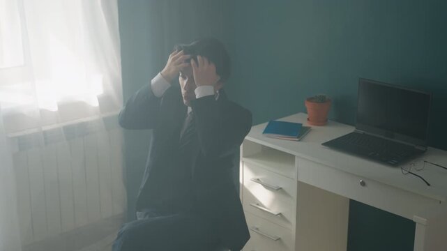Asian man in suit brooding by window desk laptop and potted plant visible, soft morning light, hand to temple, scenes show weary job applicant, junior executive, remote worker experiencing slow