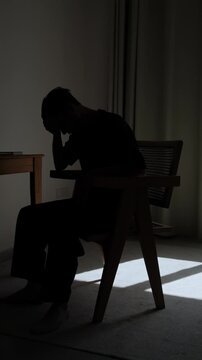 Silhouette of lonely man sitting alone in dark room, lowering his head into his hands in emotional distress, expressing sadness, despair, isolation and inner struggle, vertical