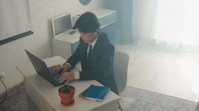 Businessman typing at laptop desk young professional in suit concentrating on screen, notebook, plant on desk, bright window light, home office, drafting report, preparing presentation, deadline