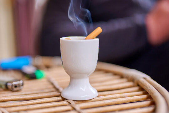 A person enjoys a cigarette placed in a white cup on a wooden table. The smoke rises into the air during a casual outdoor setting and time