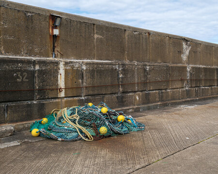Green fishing net with bright yellow floats resting on the concrete pier at Buckie Harbour in Moray Scotland during a cloudy afternoon