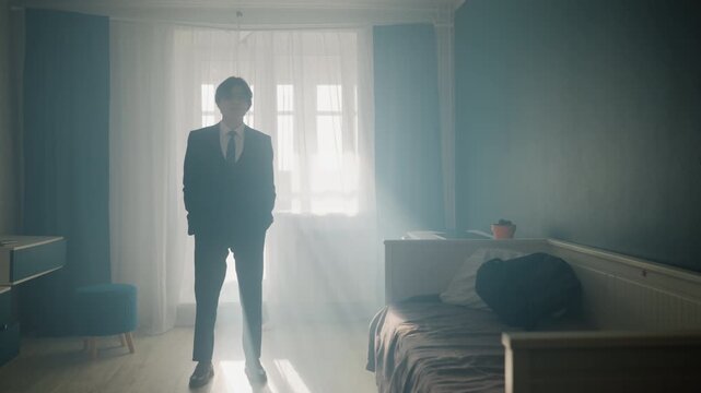 Young man in suit stands contemplative near window, morning sunlight filters through sheer curtains, soft haze fills hotel bedroom, man adjusts cufflink and pauses before departure, cinematic moody