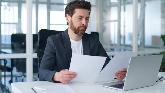 Concentrated hesitating Caucasian mature business man reading papers documents analyzing job project problem thinking businessman think holding documents middle-aged male with laptop ponder in office