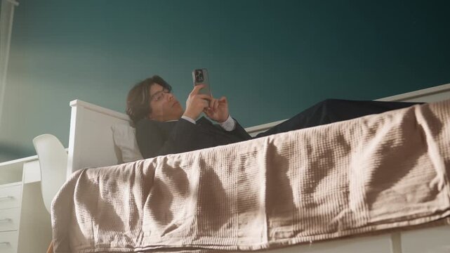 Asian man reclining on bed, scrolling through phone while planning content lowangle bedroom shot with quilted blanket foreground, teal wall and soft daylight, casual creator vibe, brainstorming