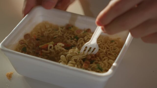 Closeup instant noodles in foam container, student uses plastic fork to stir and lift steaming ramen, visible vegetables and seasoning granules, casual indoor light, crumbs on table, quick budget