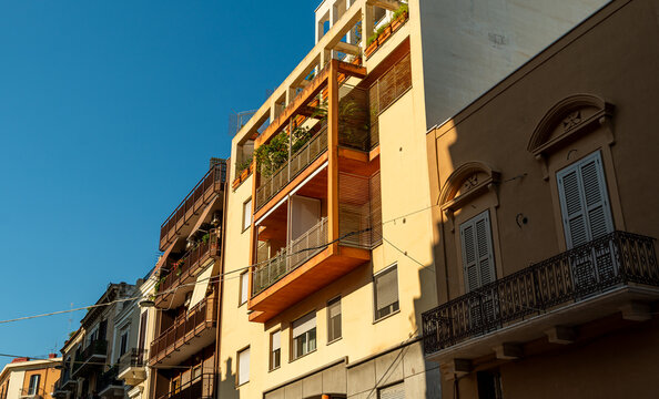 Residential building facade in Bari, Italy featuring modern wooden balconies and green plants on upper floors alongside classic Italian architecture under clear blue sky