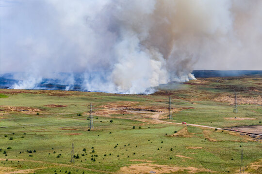 High angle view of wildfire burning across rural moorland terrain, smoke billowing past electricity pylons during dry summer heatwaves where fire risk increases