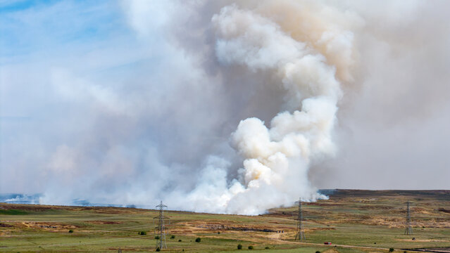 Large grass fire billowing heavy smoke clouds across dry summer terrain, electricity pylons and transmission lines crossing open ground below sky