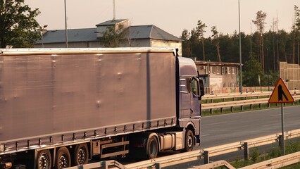 Samochód ciężarowy z szarą naczepą typu plandeka porusza się trasą szybkiego ruchu. Przy drodze widoczny jest znak ostrzegający o zwężeniu jezdni oraz infrastruktura przemysłowa w tle.