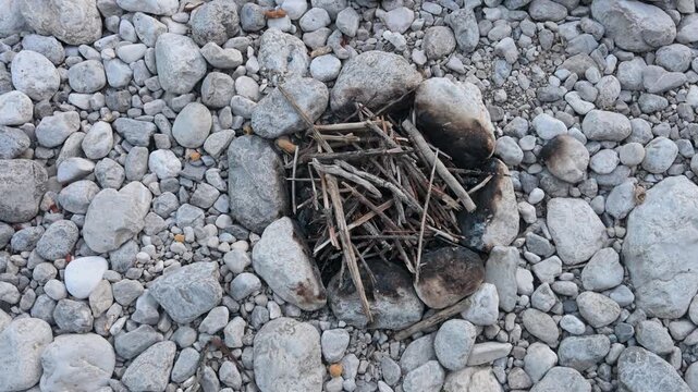 Small rustic campfire pit built with river rocks on a pebble bank. Dry twigs and branches arranged inside a stone fire ring for camping.