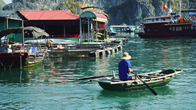 An adult man wearing a conical hat paddles a wooden rowboat in a busy floating village along a waterway on Cat Ba Island, Vietnam, with karst mountains in the background.