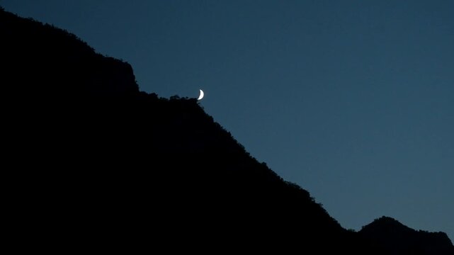 Thin crescent moon shining in a dim twilight sky above a mountainous ridge. Bright crescent moon glowing over a dark mountain slope at dusk.