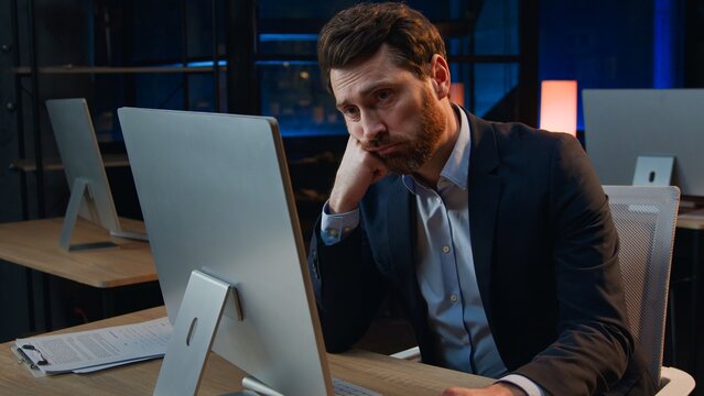 Bored unmotivated Caucasian business man working with boring computer work at night office at evening sad frustrated stressed overworked middle-aged businessman male employer feeling burnout deadline