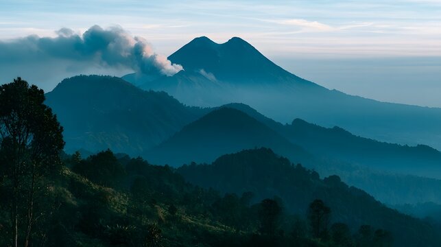 A majestic mountain range with a smoking peak rises above layered hills under a soft blue sky