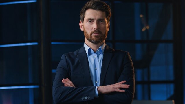 Portrait serious confident adult Caucasian business man crossing hands posing at night evening office middle aged businessman male with crossed arms looking at camera working overtime deadline work