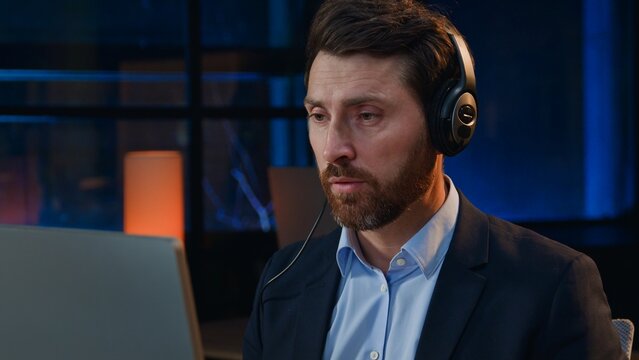 Concentrated Caucasian middle-aged business man in headphones talking video call at night evening office mature businessman male in headset consultant talk computer conference overtime helping clients