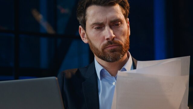 Close up serious Caucasian middle-aged business man with computer and papers at evening company businessman male corporate leader working with laptop documents reports review stats at night office
