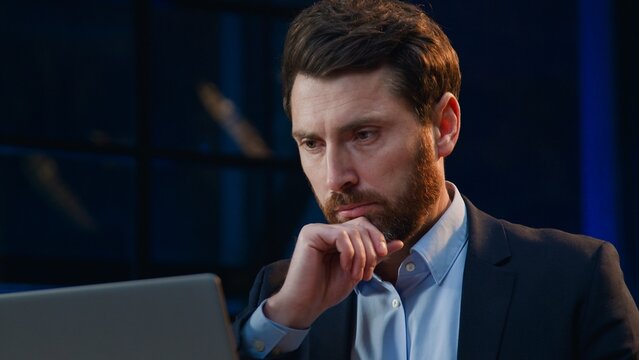 Close up troubled middle-aged Caucasian business man thinking with computer at evening company businessman male corporate leader think with laptop ponder new idea searching solution at night office