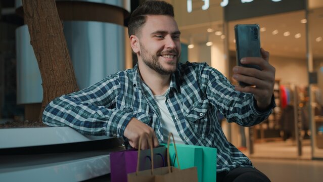 Caucasian client man with many shopping bags talking mobile phone video call recording vlog stream smiling customer with purchases smile talk smartphone conference virtual chat male guy indoor in mall